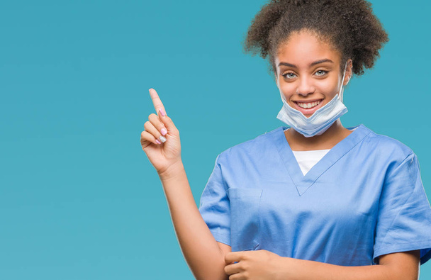 Mladý afro americký lékař žena izolované pozadí s velkým úsměvem na tváři, rukou a prstu přejdete na straně při pohledu na fotoaparát. - Fotografie, Obrázek