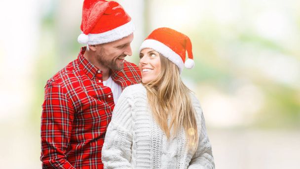 Молодая влюбленная пара в рождественской шляпе на изолированном фоне смотрит в сторону с улыбкой на лице, естественным выражением лица. Смеясь уверенно
. - Фото, изображение