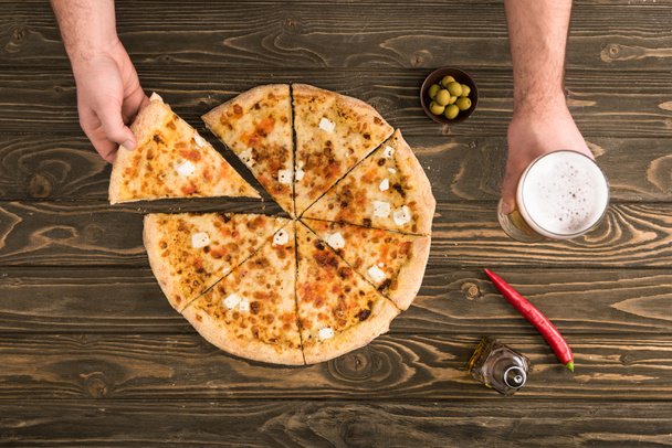 cropped view of man eating cheese pizza on wooden table with beer and olives - Photo, image