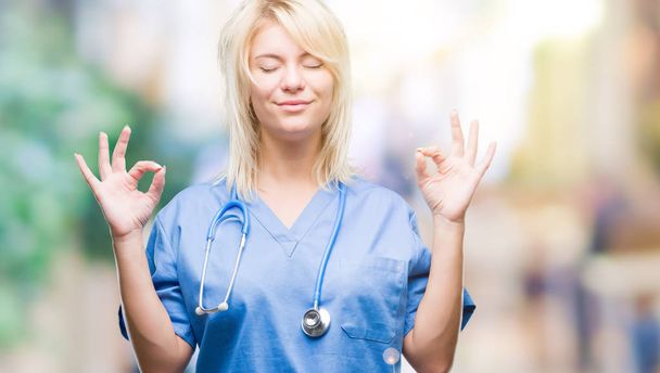 Young beautiful blonde doctor woman wearing medical uniform over isolated background relax and smiling with eyes closed doing meditation gesture with fingers. Yoga concept. - Photo, Image