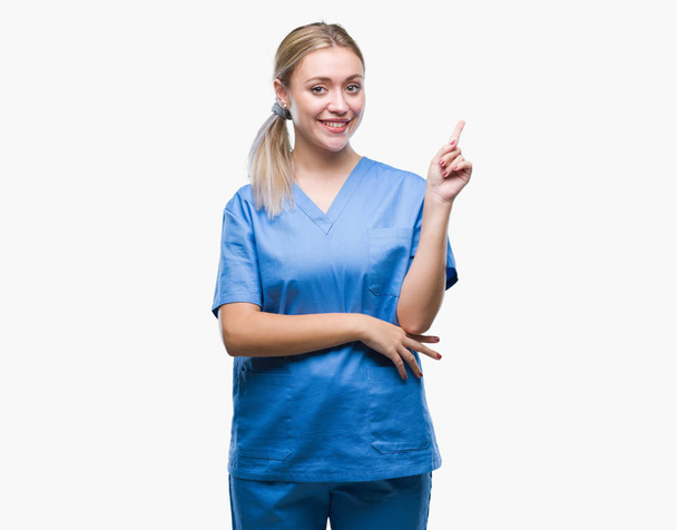 Νεαρή ξανθιά χειρουργός γιατρός γυναίκα φορώντας ιατρική στολή πάνω από απομονωμένη φόντο με ένα μεγάλο χαμόγελο στο πρόσωπο, δείχνοντας με το χέρι και το δάχτυλο στο πλάι κοιτάζοντας την κάμερα. - Φωτογραφία, εικόνα
