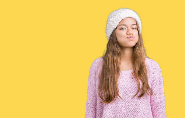 Young beautiful brunette woman wearing sweater and winter hat over isolated background puffing cheeks with funny face. Mouth inflated with air, crazy expression. - Photo, Image