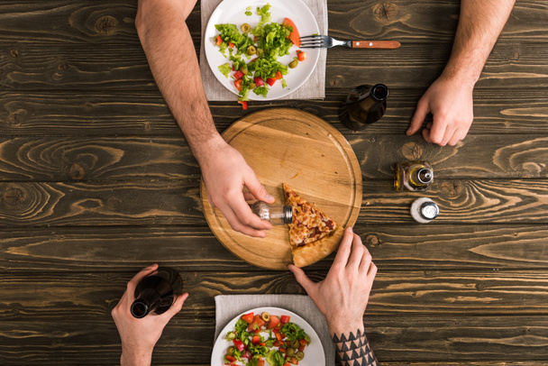 częściowy widok ludzi przyprawy kawałek pizzy, podczas posiłku w drewniany stół - Zdjęcie, obraz