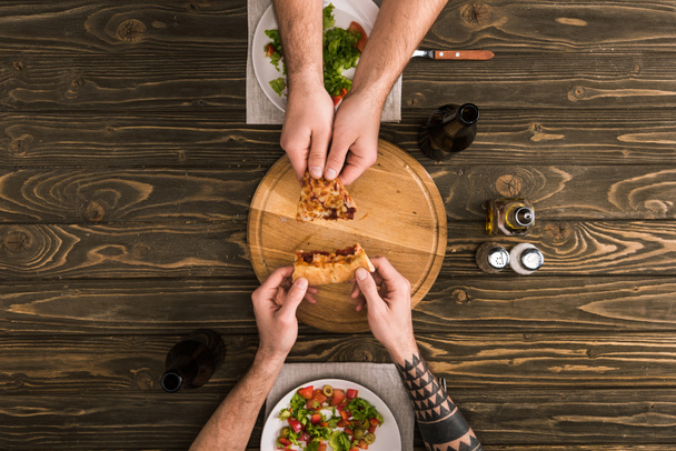 vista parziale di uomini che condividono fetta di pizza durante il pasto al tavolo di legno
 - Foto, immagini