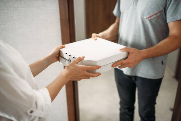 Pizza delivery man geeft kartonnen doos aan vrouwelijke klant bij de deur, het leveren van service. Courier van pizzeria en vrouw binnenshuis, afhaalmaaltijden leveren - Foto, afbeelding