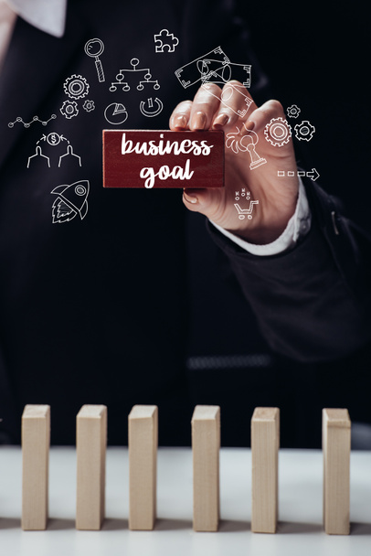 cropped view of woman holding red wooden brick with words 'business goal' isolated on black, icons on foreground - 写真・画像