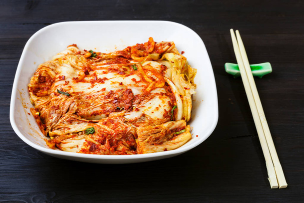 korean cuisine - kimchi appetizer (spicy nappa cabbage) in white bowl and chopsticks on dark brown wooden table - Foto, imagen