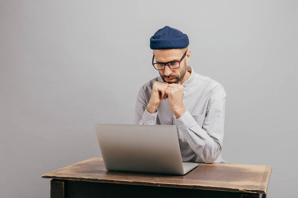 Serious male workaholic sits at desktop with modern electronic device, watches video, prepares financial report, concentrated in laptop computer, wears white shirt, optical spectacles, models indoor - 写真・画像