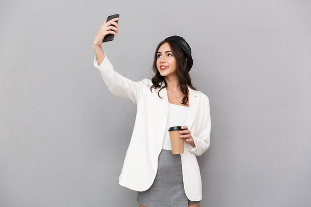 Portrait of a beautiful young woman dressed in jacket over gray background, holding cup of coffee, taking a selfie with mobile phone - Photo, image