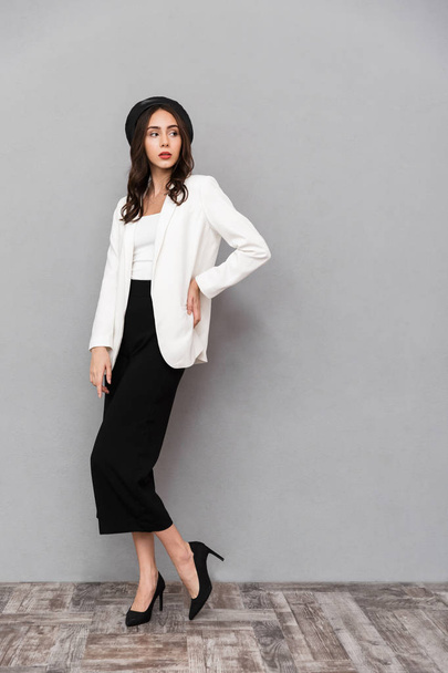 Full length portrait of a pretty young woman dressed in jacket and pants standing over gray background, looking away - Foto, imagen