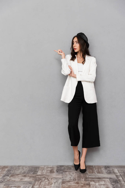 Full length portrait of a pretty young woman dressed in jacket and pants standing over gray background, pointing away - Foto, afbeelding