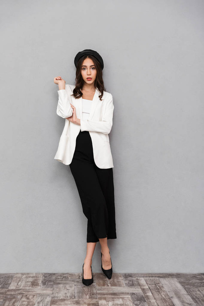 Full length portrait of a pretty young woman dressed in jacket and pants standing over gray background, looking at camera - Фото, изображение