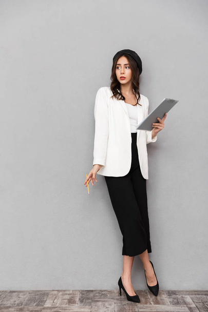 Full length portrait of a pretty young woman dressed in jacket and pants standing over gray background, holding tablet, looking away - Foto, Imagen
