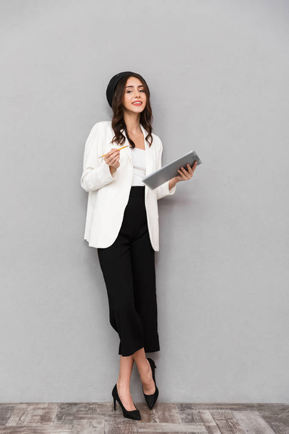 Full length portrait of a pretty young woman dressed in jacket and pants standing over gray background, holding tablet - Foto, Bild