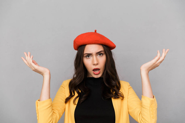 Close up of a confused young woman in red hat standing over gray background, shrugging shoulders - Foto, immagini