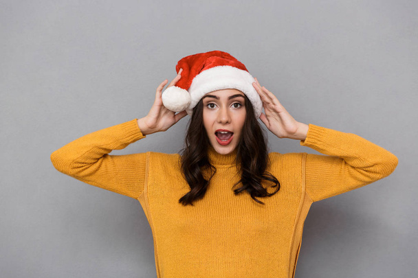 Portrait of brunette woman wearing Santa Claus red hat smiling and having fun isolated over gray background - Фото, зображення