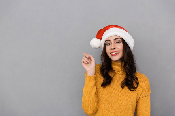Portrait of happy woman wearing Santa Claus red hat smiling and pointing finger at copyspace isolated over gray background - Fotó, kép