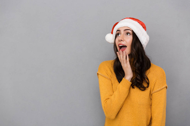 Portrait of joyful woman wearing Santa Claus red hat wondering and looking upward at copyspace isolated over gray background - Foto, afbeelding