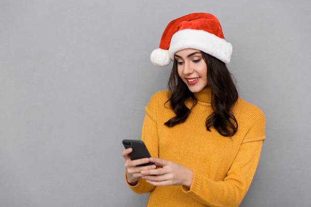 Portrait of a smiling young woman wearing red santa claus hat standing isolated over gray background, using mobile phone - 写真・画像