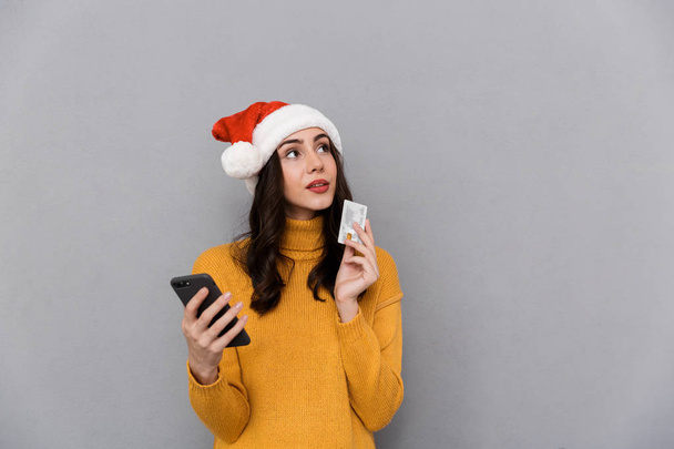 Portrait of a smiling young woman wearing red santa claus hat standing isolated over gray background, using mobile phone, showing plastic credit card - Foto, Bild