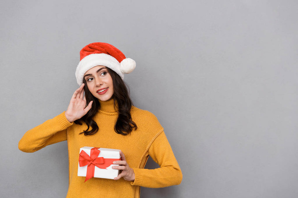 Smiling young woman wearing christmas hat standing isolated over gray background, holding present box, looking away - Fotoğraf, Görsel