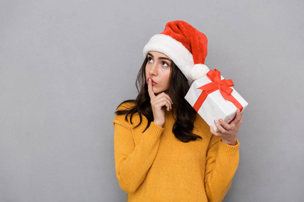 Confused young woman wearing christmas hat standing isolated over gray background, holding present box - Fotó, kép