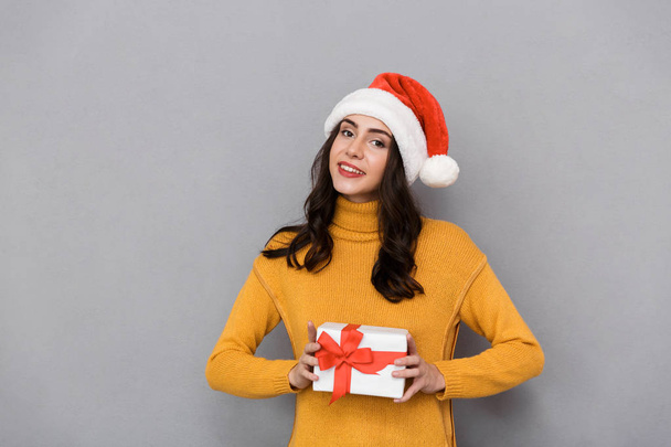 Smiling young woman wearing christmas hat standing isolated over gray background, holding present box - Foto, imagen