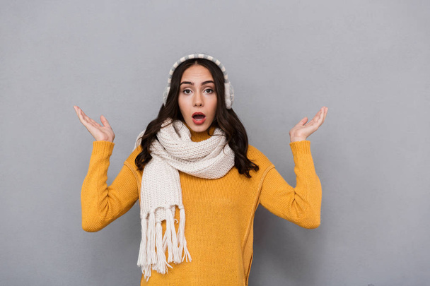 Portrait of a shocked young woman wearing sweater and scarf isolated over gray background, shrugging shoulders - Photo, image