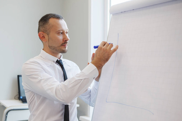 Teamwork of office staff. Businessman coach in a white shirt and tie, standing near the flipchart, explains the schedule of tasks for the future. Business training. Personal growth. - Φωτογραφία, εικόνα