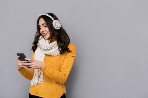 Portrait of happy woman wearing ear muffs and scarf holding mobile phone isolated over gray background - Foto, Imagem