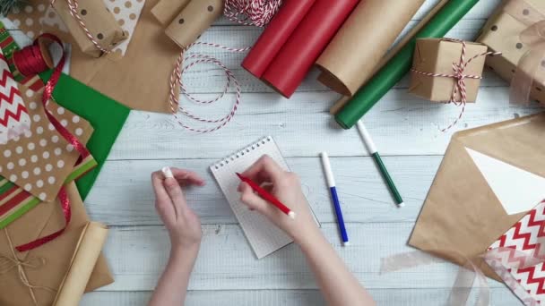 Női tini kézzel írás egy ajándék listát a télapó ajándékok fából készült asztal, csomagoló papír, piros karácsonyi díszek. Karácsonyt és boldog új évet összetétele. Lapos feküdt, top view - Felvétel, videó