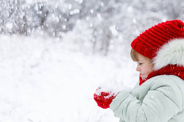 Маленька мила дівчинка, одягнена в різнокольоровий одяг, стоїть з боком на передньому плані щиро насолоджуючись купою пухнастого снігу в руці на зимовому тлі
. - Фото, зображення