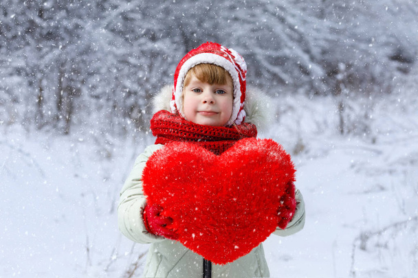 Una niña linda con ropa brillante está mostrando un enorme corazón rojo en el fondo del paisaje de invierno. El concepto es la celebración del día de San Valentín
. - Foto, Imagen