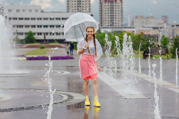 Young pretty girl with two braids in yellow boots and with transparent umbrella stands near fountain. - Valokuva, kuva