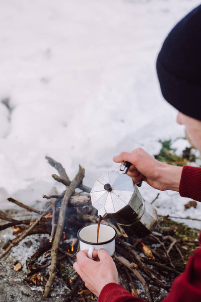 Traveling man a sits near camp fire an in winter time and pours itself hot coffee . Concept adventure active vacations outdoor. Winter camping - Photo, Image