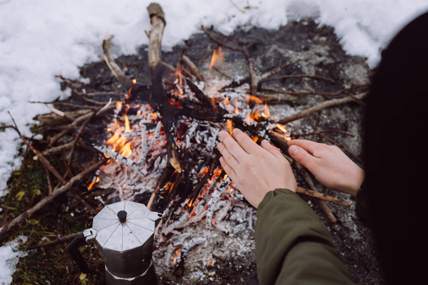 Girl in the hat warms hands near a fire in winter. Concept adventure active vacations outdoor. Winter camping - Foto, imagen