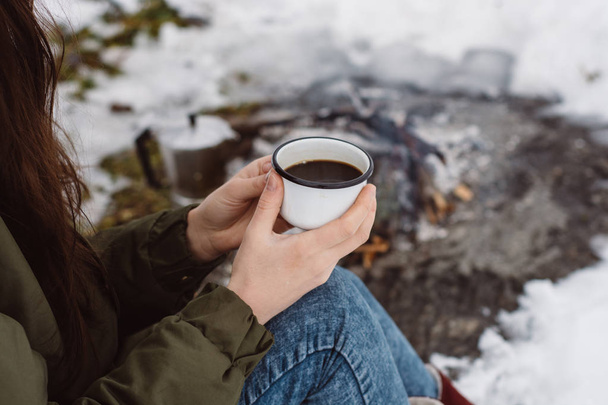 Traveling girl sits near camp fire with a cup of hot coffee an in winter time. Concept adventure active vacations outdoor. Winter camping - 写真・画像