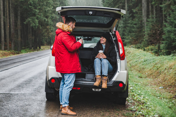 couple stop to drink hot tea. road trip concept. sitting in suv trunk at road side in mountains forest. lifestyles - Photo, Image