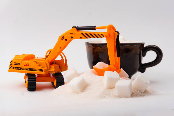 speelgoed graafmachine laadt kubus suiker in een echte kopje koffie op een lichte achtergrond - Foto, afbeelding