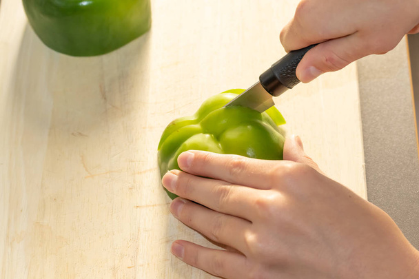 A child cuts a green sweet bell pepper on a wooden kitchen board on the kitchen table on a sunny afternoon - Foto, Imagen