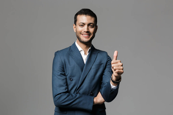 Image of positive arabic businessman 30s in formal suit looking at camera and showing thumb up isolated over gray background - Fotó, kép