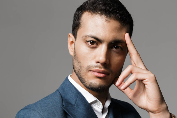 Image of concentrated arabic businessman 30s in formal suit thinking and holding index finger at forehead isolated over gray background - Foto, afbeelding