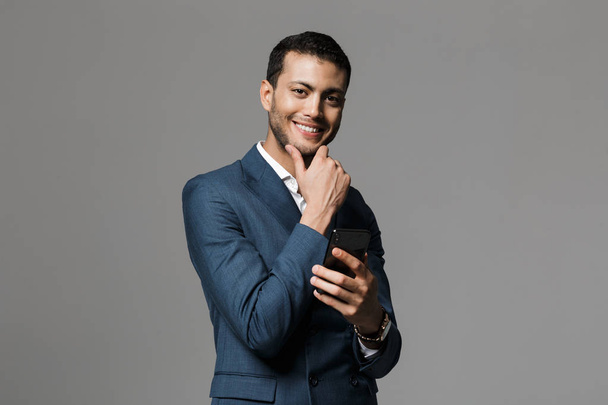 Image of a handsome young happy cheerful business man isolated over grey wall background using mobile phone. - Zdjęcie, obraz