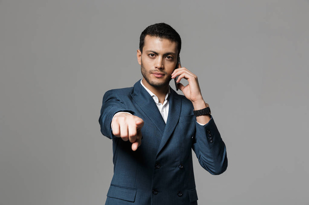 Image of handsome business man talking by mobile phone posing isolated over grey wall background pointing to you. - 写真・画像