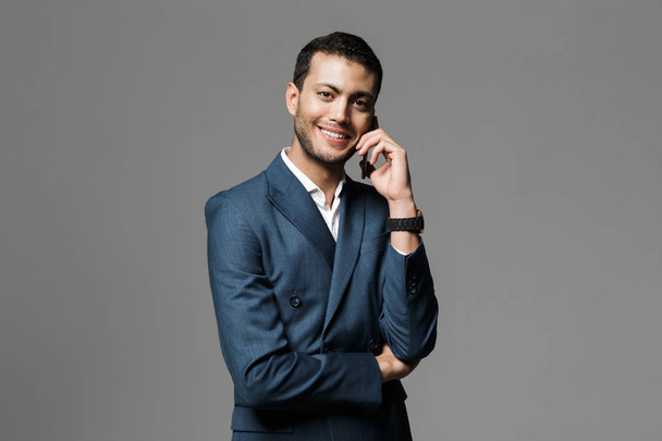 Image of a handsome happy smiling young business man isolated over grey wall background talking by mobile phone. - Zdjęcie, obraz