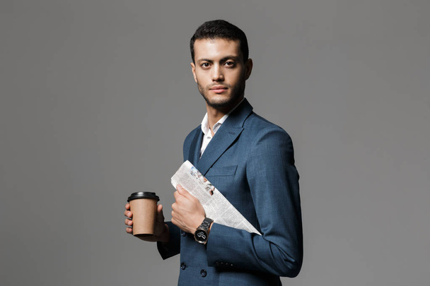 Image of confident arabic businessman 30s in formal suit holding newspaper and takeaway coffee isolated over gray background - Foto, afbeelding
