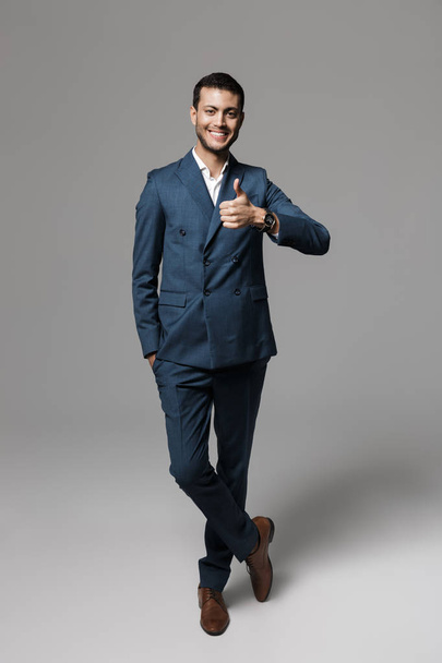 Full length image of cheerful arabic businessman 30s in formal suit gesturing at camera isolated over gray background - Foto, Bild