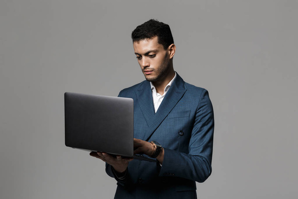 Portrait of a confident young businessman dressed in suit standing isolated over gray background, using laptop - Fotó, kép