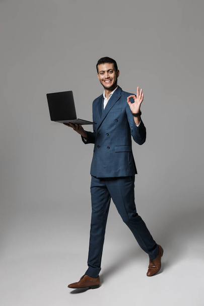 Full length photo of cheerful arabic businessman 30s in formal suit smiling and holding laptop isolated over gray background - Фото, изображение