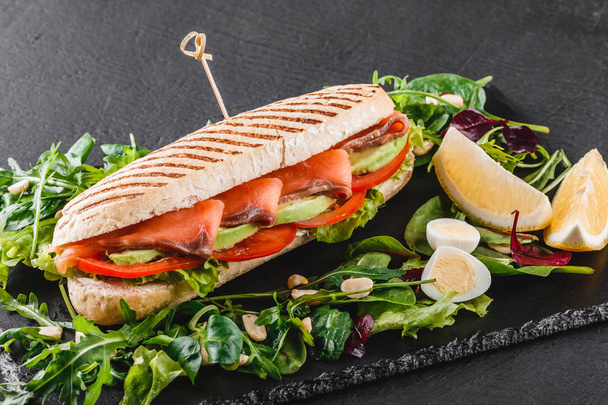 Sandwich with red fish, eggs, avocado, fresh vegetables and greens on black shale board over black stone background. Healthy food concept - Фото, зображення